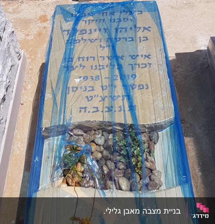 מצבה מכוסה בניילון כחול עם כיתוב בעברית ואבנים קטנות.